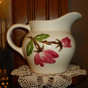 May include: White ceramic jug with a brown handle and a floral design featuring pink and green flowers. The jug is sitting on a white crocheted doily.