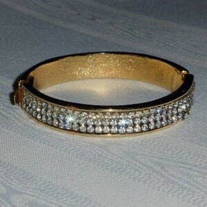 以下が含まれることがあります： 縁にクリアなラインストーンが並んだゴールドのバングルブレスレットです。