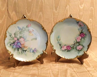 Peut inclure: Deux assiettes en porcelaine vintage avec des bordures dorées et des motifs floraux. Une assiette présente des fleurs roses et violettes, tandis que l'autre présente des roses roses.