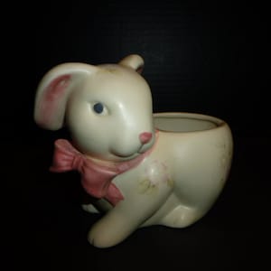 Könnte beinhalten: Ein weißer Keramik-Hasentopf mit einer rosa Schleife und Blumendetails. Der Topf hat eine kleine Öffnung oben zum Pflanzen.