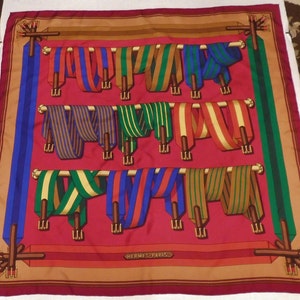 Puede incluir: Un pañuelo de seda rojo con un patrón de cintas rayadas de colores que cuelgan de un estante de madera. El pañuelo tiene un borde marrón y el texto "HERMES PARIS" en el centro.