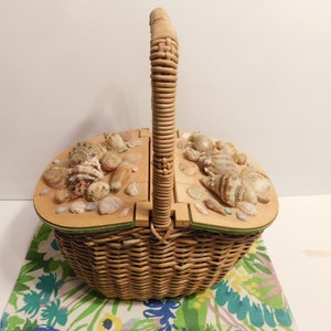 May include: A wicker picnic basket with a hinged lid decorated with seashells. The basket has a woven handle and a green trim around the top.