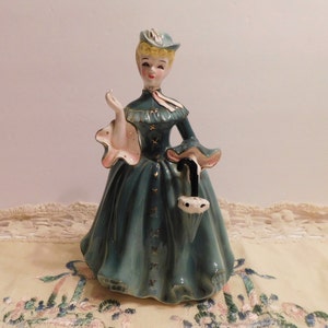 Peut inclure: Une figurine en céramique d'une femme en robe et chapeau turquoise, tenant un parapluie noir et blanc. Elle a les cheveux blonds et un sourire peint.