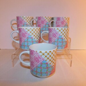 May include: A set of six white ceramic mugs with a colorful patchwork design. The design includes pink floral patterns, blue geometric shapes, and a gold trellis pattern. Each mug has a white handle.