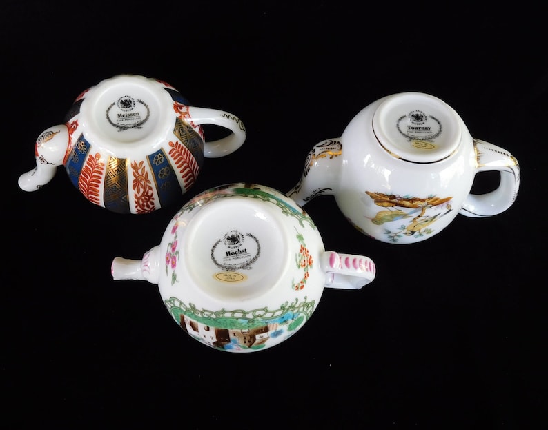 May include: Three decorative porcelain teapots with intricate designs. One features a pattern of blue, red, and gold stripes. Another has a landscape scene with birds, and the third displays a floral and architectural motif. Each teapot has a handle and spout.