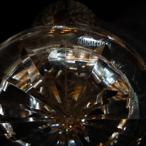 May include: A clear glass bottle stopper with a cut-glass design. The stopper has a round base and a pointed top. The glass is clear and reflects light.