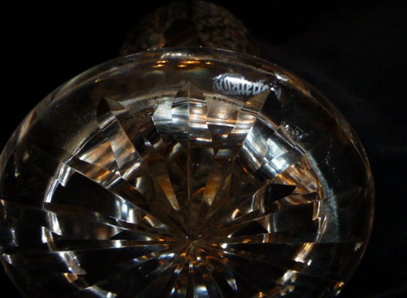 May include: A clear glass bottle stopper with a cut-glass design. The stopper has a round base and a pointed top. The glass is clear and reflects light.