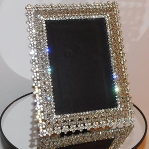 Pode incluir: Uma moldura retangular com interior preto e borda prateada incrustada de joias. A moldura é exibida em uma superfície espelhada, refletindo seus detalhes brilhantes. O design da moldura apresenta um padrão floral.