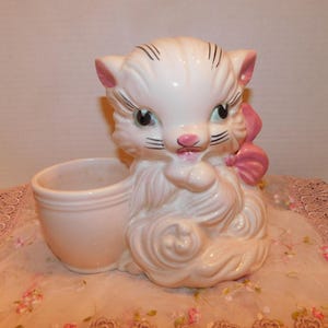 May include: A white ceramic planter shaped like a cat with a pink bow. The cat has black eyes and whiskers and a pink nose. The planter has a separate pot for planting.
