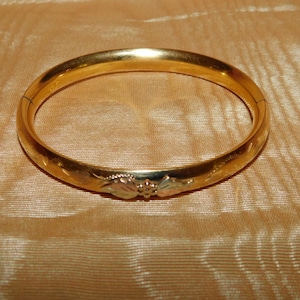 May include: A gold-colored bangle bracelet with floral detailing. The bracelet is circular and has a clasp. The surface of the bracelet has etched floral patterns. The background is a gold-colored fabric.