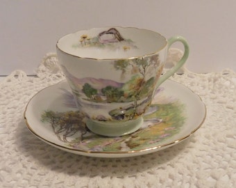 May include: A vintage teacup and saucer set with a green and white floral design. The cup and saucer feature a hand-painted landscape scene with a cottage and trees.