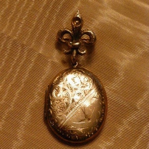May include: A gold locket with a fleur-de-lis design on the top and an engraved floral pattern on the front.