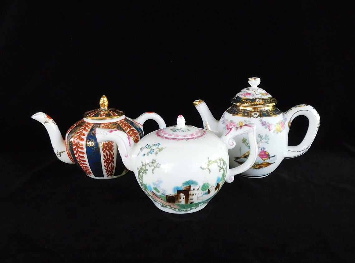 May include: Three decorative teapots with intricate designs. The teapots feature a variety of patterns, including floral, striped, and landscape scenes. The teapots are white with gold accents and colorful details. The teapots are displayed against a black background.