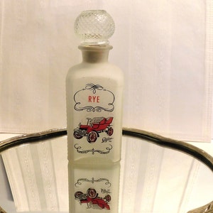 May include: White frosted glass decanter with a clear glass stopper. The decanter has a red and black image of a vintage Cadillac car with the word "RYE" in red lettering on the front.
