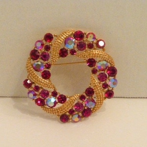 May include: A gold tone brooch with a circular design featuring a swirl of pink and iridescent rhinestones.