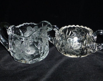 Op de afbeelding: Twee glazen serveerschalen, een melkkan en een suikerpot, met geëtste bloemmotieven. De melkkan heeft een handvat en een schenktuit, terwijl de suikerpot twee handvatten heeft. Beide hebben geschulpte randen, tegen een zwarte achtergrond.