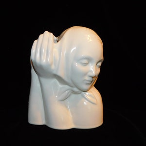May include: White ceramic sculpture of a woman's head and shoulders, with her head tilted down and her hand resting on her chin. The sculpture is a vase with a smooth, glossy finish.