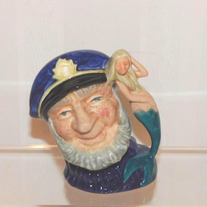 May include: A ceramic character jug depicting a bearded sailor wearing a dark blue captain's hat with a gold emblem. A small mermaid with long blonde hair and a teal tail is perched on the side of the hat, serving as the handle for the vessel.