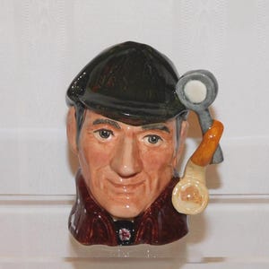 May include: A ceramic character jug depicting a person wearing a dark green deerstalker hat and a brown coat. The figure holds a light-coloured pipe with a grey bowl near the right side of the face. The facial features are painted with realistic skin tones.