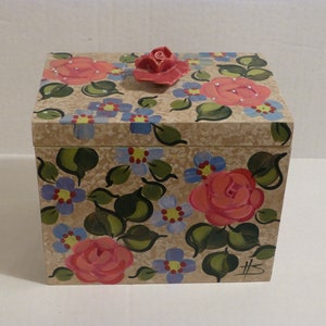 May include: A square wooden box with a floral design painted on the lid and sides. The box has a beige background with pink roses, blue flowers, and green leaves. The lid has a small pink rose on top.
