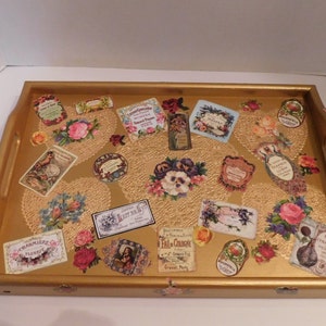 May include: A gold-colored wooden tray with a decorative design featuring vintage labels, flowers, and lace. The tray has two handles on the sides.