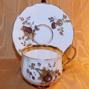 May include: A white teacup and saucer set with a gold rim and handle. The cup and saucer are decorated with brown and gold fruit and floral designs. The saucer has a scalloped edge.