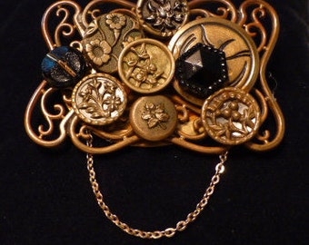 May include: A gold-toned brooch with a chain attached, featuring a variety of decorative buttons and a black glass cabochon. The buttons have floral and geometric designs.