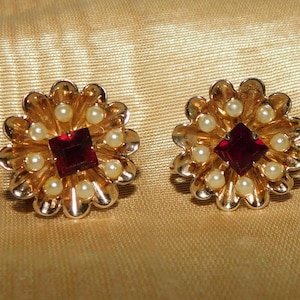 Könnte beinhalten: Ein Paar goldfarbene Ohrringe mit einem roten Juwelenmittelpunkt und einem Kreis aus weißen Perlen um das Juwel. Die Ohrringe sind blumenförmig.