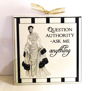 Könnte beinhalten: Quadratische dekorative Fliese mit einem schwarz-weiß gestreiften Rand. Die Fliese zeigt eine Vintage-Illustration einer Frau in einem Kleid mit dem Text "QUESTION AUTHORITY - ASK ME anything" in schwarzer Schrift.