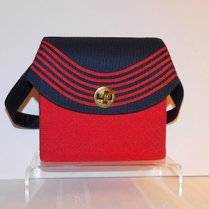 May include: A small, rectangular red handbag with a navy blue flap featuring three red stripes. The bag has a gold-coloured clasp and a black strap. The bag is displayed on a clear stand.