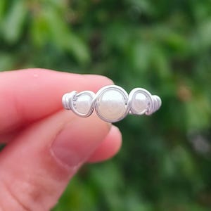 Könnte beinhalten: Ein handgefertigter Ring aus silbernem Draht mit drei Kunstperlen. Das Design des Rings zeigt eine zentrale, größere Perle, flankiert von zwei kleineren Perlen, alle in einem zarten, gewebten Drahtrahmen. Der Ring wird in einer Hand gehalten.