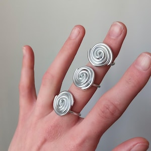 Op de afbeelding: Drie zilverkleurige ringen met een spiraalvormig ontwerp worden op vingers getoond. De ringen hebben een eenvoudige, moderne uitstraling. Het spiraalontwerp is het middelpunt en zorgt voor een unieke en opvallende look. De ringen staan tegen een neutrale achtergrond.
