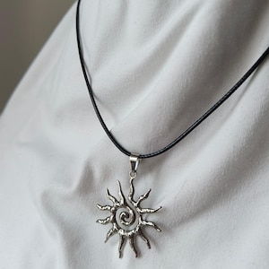 Pode incluir: Colar com pingente de sol prateado e cordão preto. O pingente apresenta um centro em espiral e raios radiantes. O colar é exibido em um fundo branco.