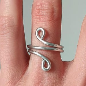 Op de afbeelding: Een zilverkleurige draadring met een wervelend ontwerp. De ring is om een vinger gewikkeld en toont zijn unieke, handgemaakte stijl. Het ontwerp van de ring heeft twee lussen en een spiraal, wat een elegante en artistieke uitstraling creëert.