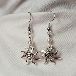 Op de afbeelding: Zilverkleurige oorbellen met een zonmotief, een spiraalvormig centrum en een hangende parel. De oorbellen hebben een haaksluiting en worden getoond tegen een witte achtergrond.