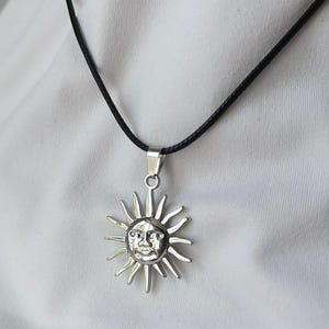 Op de afbeelding: Zilverkleurige zonnehangerketting met een zwart koord. De zonnehanger heeft een lachend gezicht en stralende stralen. De ketting wordt getoond tegen een witte achtergrond.