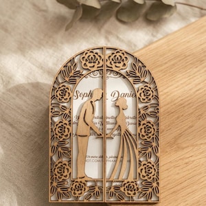 Peut inclure: Un faire-part de mariage en bois en forme d'arche avec des motifs floraux découpés au laser et les silhouettes d'une personne en costume et d'une personne en robe. Le centre affiche du texte imprimé avec les prénoms Sophia et Daniel.