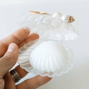 Puede incluir: Un recipiente de plástico blanco translúcido con forma de concha que se abre mediante una bisagra. El exterior superior de la concha está decorado con una pequeña estrella de mar dorada, una concha blanca y una perla blanca fijada.