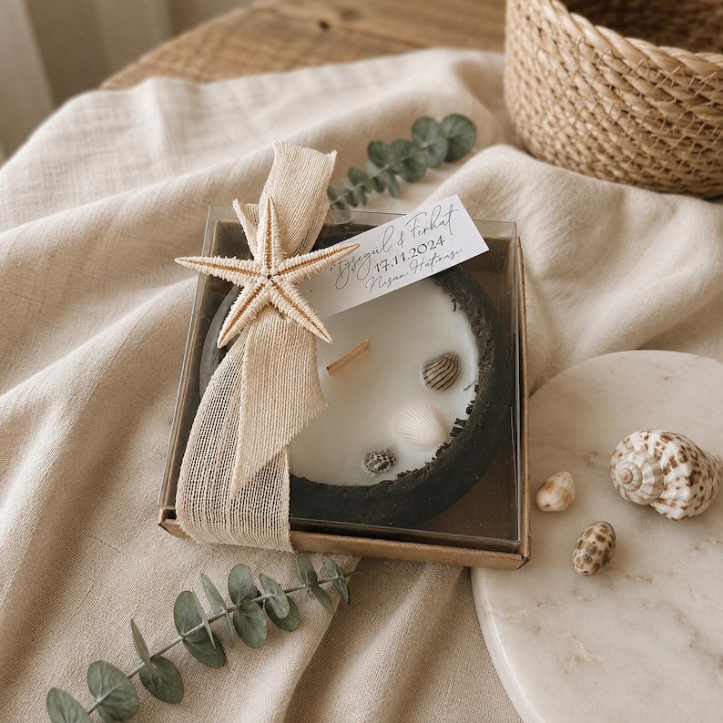 Puede incluir: Una vela redonda en un recipiente de hormig&oacute;n gris oscuro, rellena de cera blanca y peque&ntilde;as conchas marinas. Est&aacute; empaquetada en una caja transparente con una cinta beige, una estrella de mar y una etiqueta blanca con el texto Aysegul & Ferhat 17.11.2024 Nisan Hatirasi.