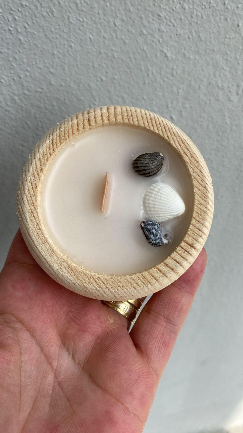 Puede incluir: Una vela redonda en un cuenco de madera clara con cera blanca lisa, una mecha de madera vertical y tres peque&ntilde;as conchas marinas incrustadas en la superficie, incluyendo una concha oscura rayada, una blanca y una moteada.