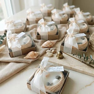 Puede incluir: Varios cuencos de madera redondos con velas de cera blanca, decorados con peque&ntilde;as conchas marinas y una estrella de mar. Las velas est&aacute;n en cajas de cart&oacute;n marr&oacute;n cuadradas con tapas transparentes, atadas con cinta blanca y una etiqueta con texto.