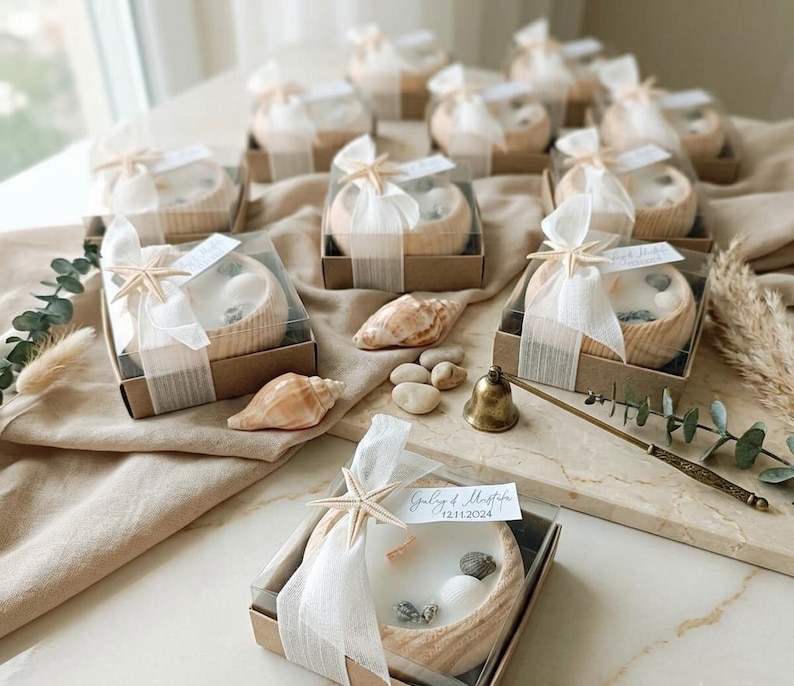 Puede incluir: Varios cuencos de madera redondos con velas de cera blanca, decorados con peque&ntilde;as conchas marinas y una estrella de mar. Las velas est&aacute;n en cajas de cart&oacute;n marr&oacute;n cuadradas con tapas transparentes, atadas con cinta blanca y una etiqueta con texto.