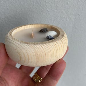 Puede incluir: Un cuenco redondo de madera natural clara que contiene cera blanca con una mecha de madera. Incrustadas en la cera hay tres peque&ntilde;as conchas marinas distintas, incluida una concha blanca y dos conchas m&aacute;s peque&ntilde;as, oscuras y estampadas.