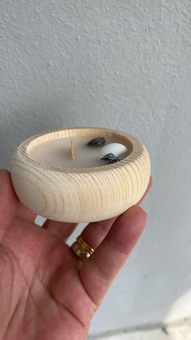 Puede incluir: Un cuenco redondo de madera natural clara que contiene cera blanca con una mecha de madera. Incrustadas en la cera hay tres peque&ntilde;as conchas marinas distintas, incluida una concha blanca y dos conchas m&aacute;s peque&ntilde;as, oscuras y estampadas.