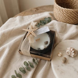 Puede incluir: Una caja de regalo cuadrada que contiene una vela blanca con mecha de madera, decorada con una estrella de mar beige, una cinta marr&oacute;n claro y conchas peque&ntilde;as. Una etiqueta blanca lleva el texto Aysegul & Ferhat 17.11.2024 Nisan Hatirasi.