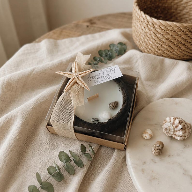 Puede incluir: Una caja de regalo cuadrada que contiene una vela blanca con mecha de madera, decorada con una estrella de mar beige, una cinta marr&oacute;n claro y conchas peque&ntilde;as. Una etiqueta blanca lleva el texto Aysegul & Ferhat 17.11.2024 Nisan Hatirasi.