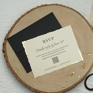 May include: An off-white RSVP card with deckled edges and gold foil accents, featuring black text and a QR code. The card rests on a black envelope, accompanied by a gold ring in a small box, a loose gold ring, and black metal scissors.