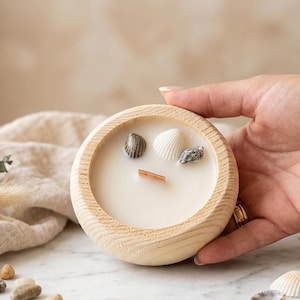 Puede incluir: Un cuenco redondo de madera clara que contiene cera de vela blanca. La vela presenta una mecha de madera plana y rectangular centrada en la cera, rodeada por tres peque&ntilde;as conchas marinas naturales dispuestas sobre la superficie de la cera.