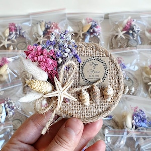 Könnte beinhalten: Ein runder Hochzeitsgastgeschenk aus Jute mit Seestern, Muscheln und Trockenblumen in Rosa, Lila und Weiß. Das Gastgeschenk hat ein kleines Etikett mit Namen und Datum.