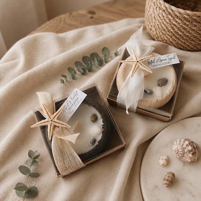 Puede incluir: Dos velas redondas en cajas cuadradas, una con base gris oscuro y otra con base de madera clara. Cada vela contiene cera blanca, peque&ntilde;as conchas, una estrella de mar beige, una cinta blanca y una etiqueta con nombres y fechas.
